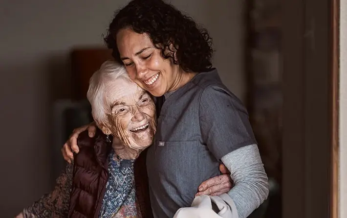 Eine Pflegekraft in Arbeitskleidung umarmt eine ältere Frau im Türrahmen. Beide lächeln.