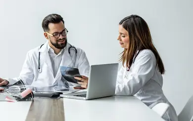 Das Bild zeigt zwei Ärztinnen und einen Arzt bei einer Besprechung. Sie sitzen an einem Tisch und diskutieren. Vor ihnen steht ein aufgeklappter Laptop.