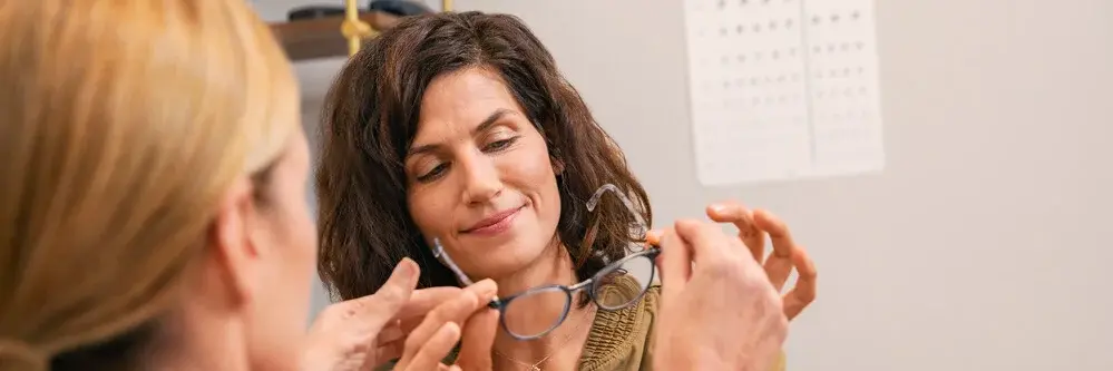 Augenoptikerin überreicht einer Frau eine Brille zur Anprobe, im Hintergrund ist eine Sehtafel sichtbar.