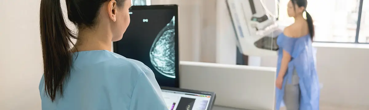 Eine Arzthelferin schaut auf einen Monitor, auf dem das Röntgenbild einer Mammographie zu sehen ist. Im Hintergrund steht eine Patienten vor dem Röntgengerät. 