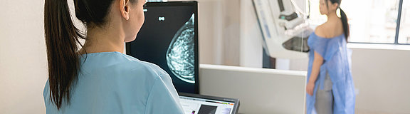 Eine Arzthelferin schaut auf einen Monitor, auf dem das Röntgenbild einer Mammographie zu sehen ist. Im Hintergrund steht eine Patienten vor dem Röntgengerät. 