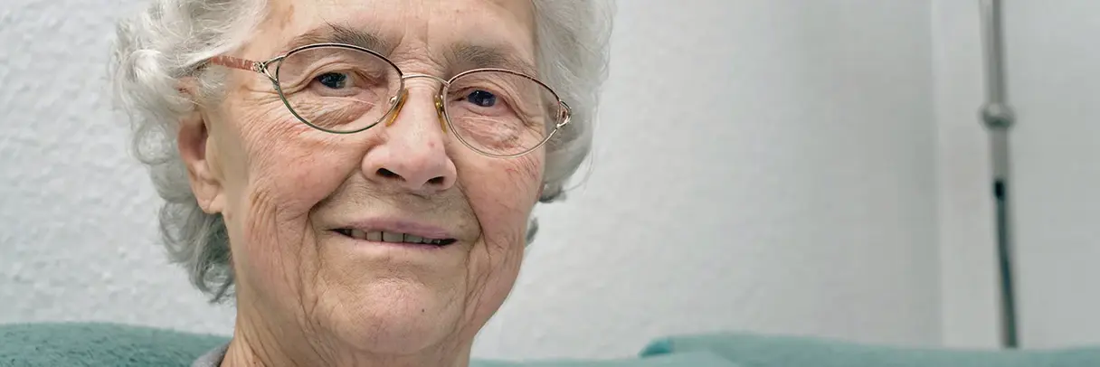 Ältere Frau mit kurzen weißen Haaren und Brille lächelt leicht, sitzt vor einer weißen Wand