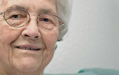 Ältere Frau mit kurzen weißen Haaren und Brille lächelt leicht, sitzt vor einer weißen Wand
