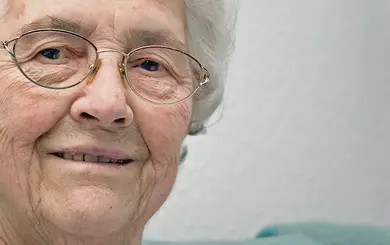 Ältere Frau mit kurzen weißen Haaren und Brille lächelt leicht, sitzt vor einer weißen Wand