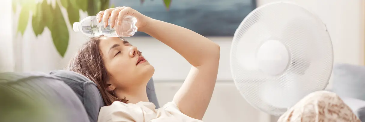 Frau sitzt auf Sofa, hält Wasserflasche an Stirn, neben ihr steht ein Ventilator, im Hintergrund Pflanzen und Fenster. Das Bild illustriert einen Beitrag zum QISA-Band F3 „Gesundheitsversorgung im Klimawandel“.