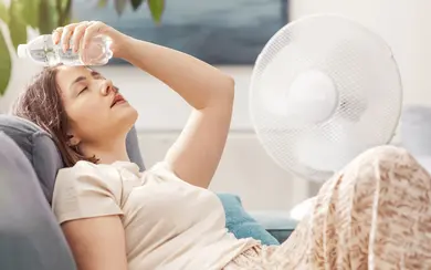 Frau sitzt auf Sofa, hält Wasserflasche an Stirn, neben ihr steht ein Ventilator, im Hintergrund Pflanzen und Fenster. Das Bild illustriert einen Beitrag zum QISA-Band F3 „Gesundheitsversorgung im Klimawandel“.