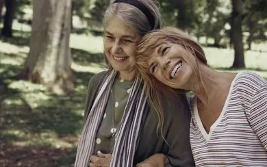 Zwei Frauen im Alter von 60 bis 70 Jahren. Eine stützt die andere. Beide lachen.