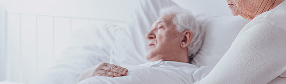 Ältere Frau unterstütz kranken Mann der im einem Krankenhausbett liegt