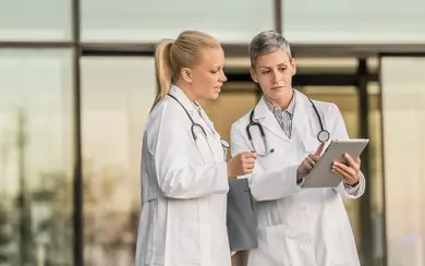 Zwei Ärztinnen in weißen Kitteln diskutieren. Die Ärztin auf der rechten Seite hält ein Tablet, während die Ärztin auf der linken Seite ein Tablet in der Hand hat. Beide tragen Stethoskope um den Hals.