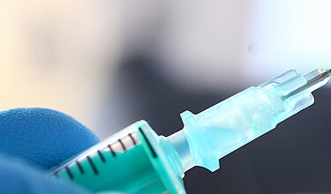 Hand mit in blauem Schutzhandschuhe hält Spritze mit Flüssigkeit (Symbolbild)