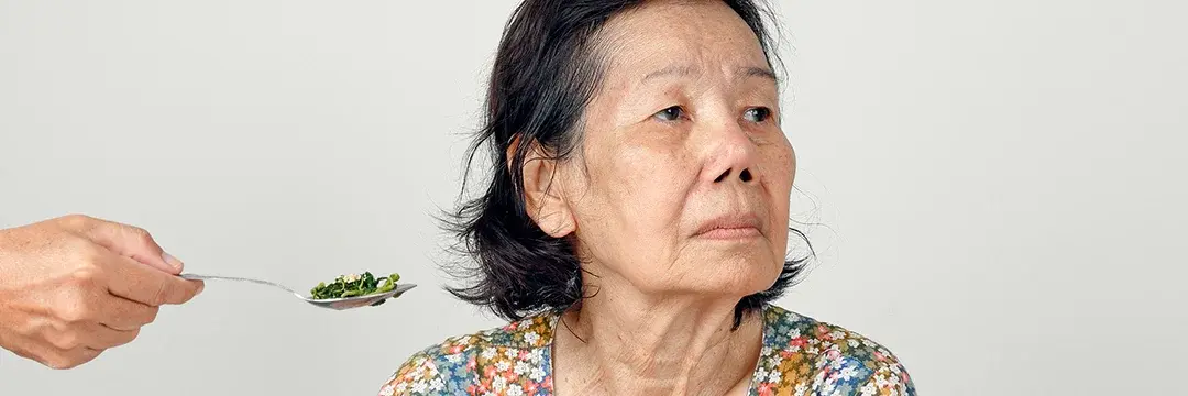 Älter Frau dreht den Kopf weg. Ihr wird ein Löffel gereicht