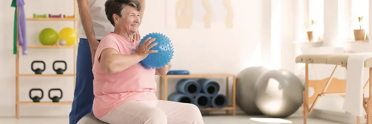 Seniorin sitzt auf einem Gymnastikball, hält einen Ball in beiden Händen und wird von einer Fachkraft gestützt