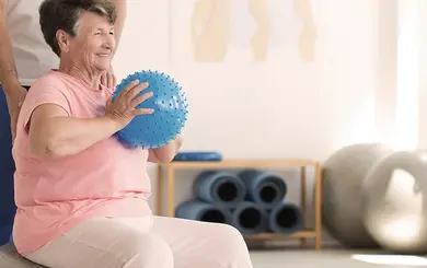 Seniorin sitzt auf einem Gymnastikball, hält einen Ball in beiden Händen und wird von einer Fachkraft gestützt