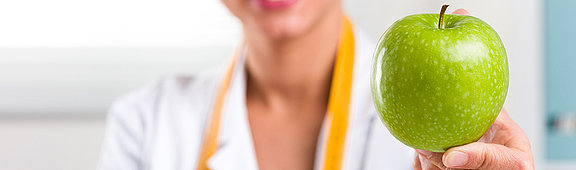 Junge Frau mit weißen Kittel und Maßband um ihren Hals, hält einen grünen Apfel 