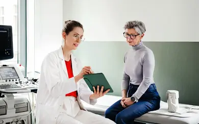 Junge Ärztin spricht mit einer älteren Patienten. Zusammen schauen sie sich Informationen auf einen Tablet-PC an
