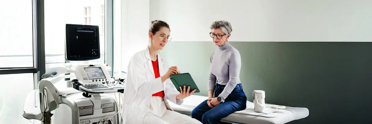 Junge Ärztin spricht mit einer älteren Patienten. Zusammen schauen sie sich Informationen auf einen Tablet-PC an