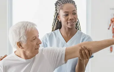 Das Bild zeigt eine Physiotherapeutin, die einen älteren Mann mit Schulterbeschwerden behandelt.