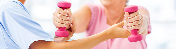 Junge weibliche Therapeutin hilft einer älteren Frau die zwei rosa Hanteln in der Hand hält 
