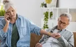 Foto: Frau und Mann auf einem Sofa – während der Mann sich ans Herz greift, ruft die Frau telefonisch nach Hilfe.