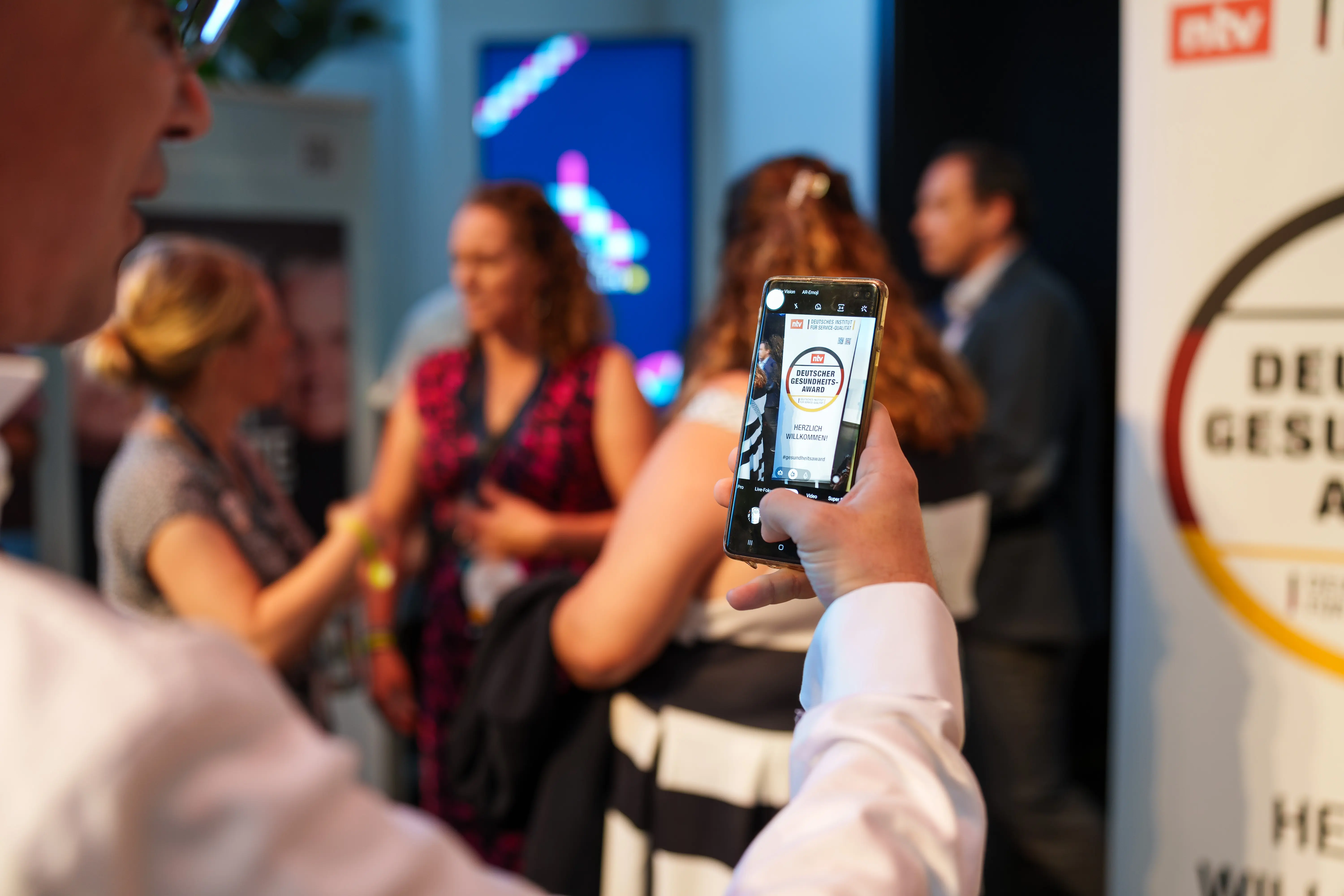 Das Bild zeigt Gäste des Deutschen Gesundheits-Award 2023, im Vordergrund ein Handy, das das Veranstaltungsposter fotografiert. Bei der Veranstaltung wurden die AOK-Angebote Gesundheitsnavigator und Pflegenavigator ausgezeichnet.