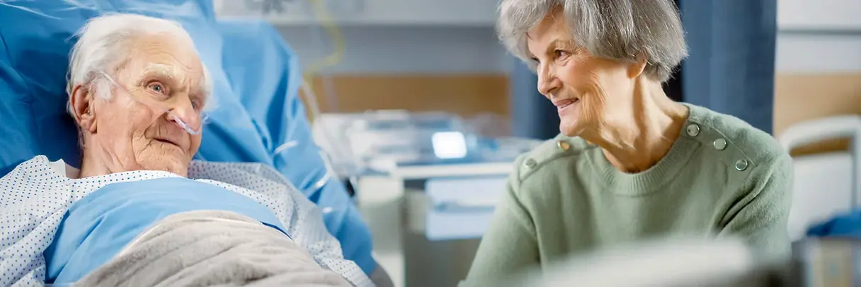 Hochbetagter Mensch im Krankenhaus Hochbetagter Mann im Krankenhausbett mit Sauerstoffbrille, lächelnde ältere Frau sitzt neben ihm.