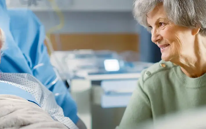 Hochbetagter Mann im Krankenhausbett mit Sauerstoffbrille, lächelnde ältere Frau sitzt neben ihm.