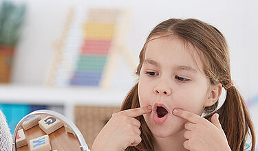 Therapeutin sitzt mit jungen Mädchen vor einem Tischspiegel und halten sich die Finger an den geöffneten Mund
