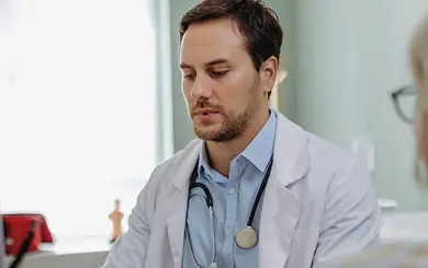 Ein Arzt mit weißem Kittel und Stethoskop um den Hals sitzt an einem Schreibtisch vor einem Laptop. Vor ihm sitzt eine Patientin mit blonden Haaren und Brille, die von hinten zu sehen ist. Die Patientin bespricht mit dem Arzt ihren Wunsch nach Einsicht in ihre Krankenakte.
