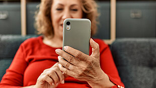Frau mittleren Alters sitzt mit Smartphone auf dem Sofa (Symbolbild)