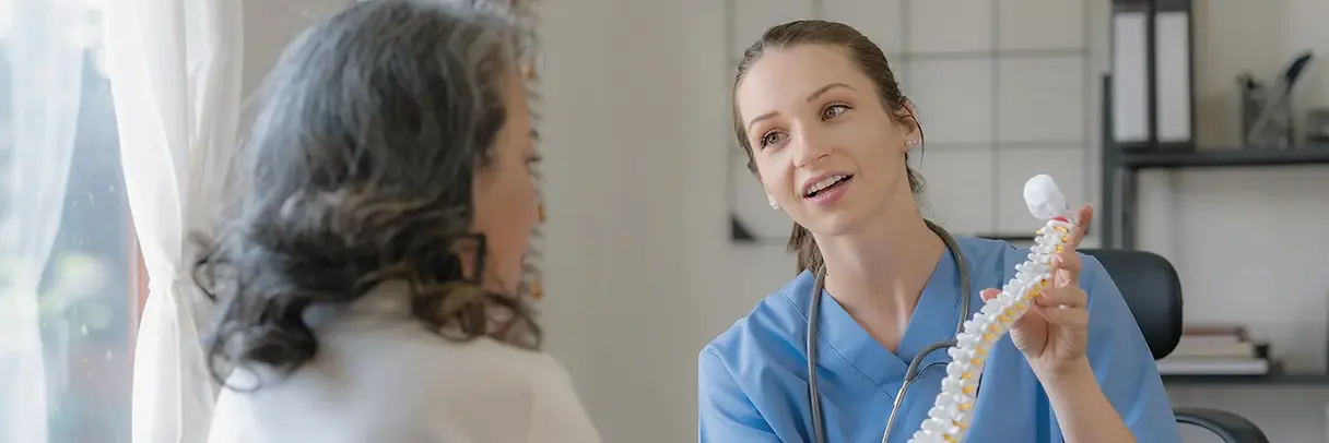 Junge Ärztin erklärt einer Patientin anschaulich ein Wirbelsäulenmodell und erläutert dabei freundlich die Zusammenhänge.