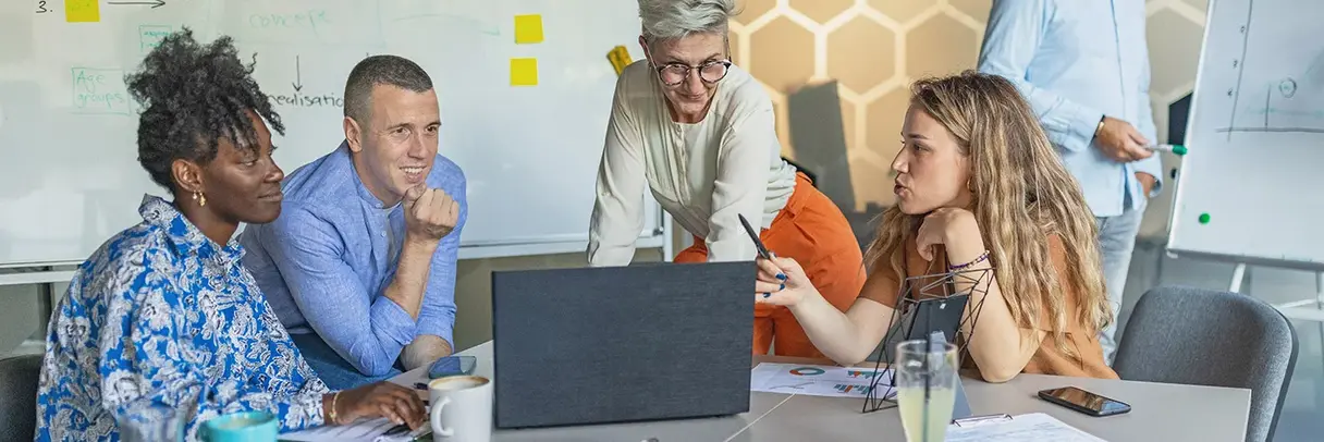 Das Bild zeigt vier Personen bei der gemeinsamen Arbeit an einem Projekt. Sie schauen gemeinsam auf einen Laptop; im Hintergrund sieht man Notizen auf bunten Post-it-Zetteln.