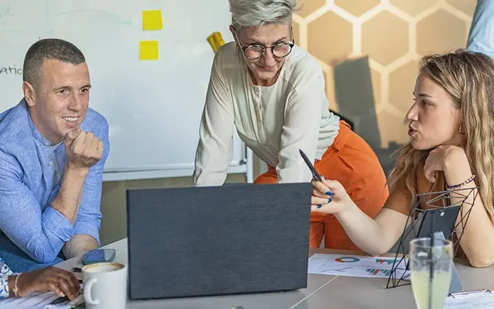 Das Bild zeigt vier Personen bei der gemeinsamen Arbeit an einem Projekt. Sie schauen gemeinsam auf einen Laptop; im Hintergrund sieht man Notizen auf bunten Post-it-Zetteln.
