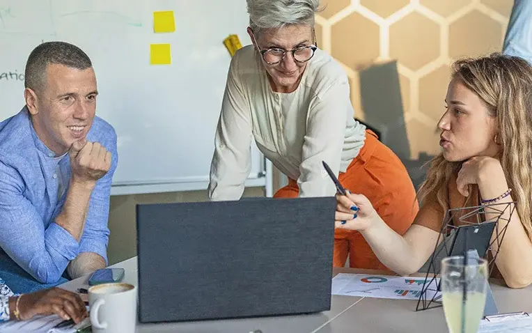 Das Bild zeigt vier Personen bei der gemeinsamen Arbeit an einem Projekt. Sie schauen gemeinsam auf einen Laptop; im Hintergrund sieht man Notizen auf bunten Post-it-Zetteln.