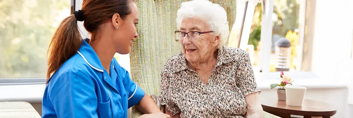 Pflegekraft mit Seniorin Eine Pflegerin in blauer Berufskleidung sitzt lächelnd neben einer älteren Frau mit Brille, die in einem gemusterten Hemd gekleidet ist. Die beiden unterhalten sich freundlich in einer hellen, gemütlichen Umgebung mit einem Sessel und einem Tisch mit Blumen im Hintergrund.