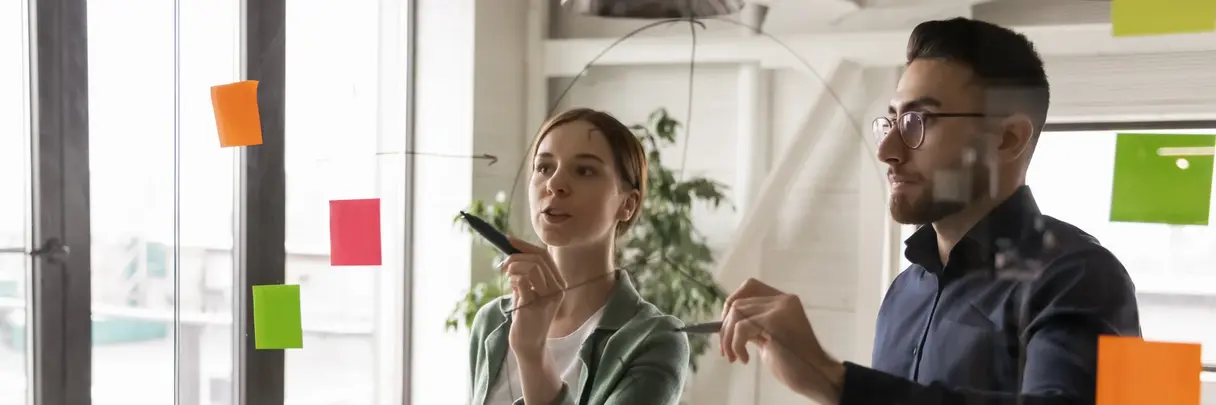 Zwei junge Personen diskutieren an einer Glaswand über Workflows diskutiert oder Arbeitsprojektprozesse: Symbolbild für Innovation und agiles Arbeiten