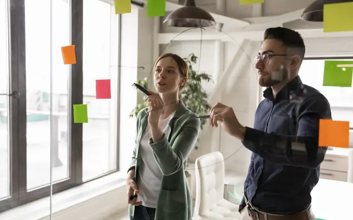 Zwei junge Personen diskutieren an einer Glaswand über Workflows diskutiert oder Arbeitsprojektprozesse: Symbolbild für Innovation und agiles Arbeiten
