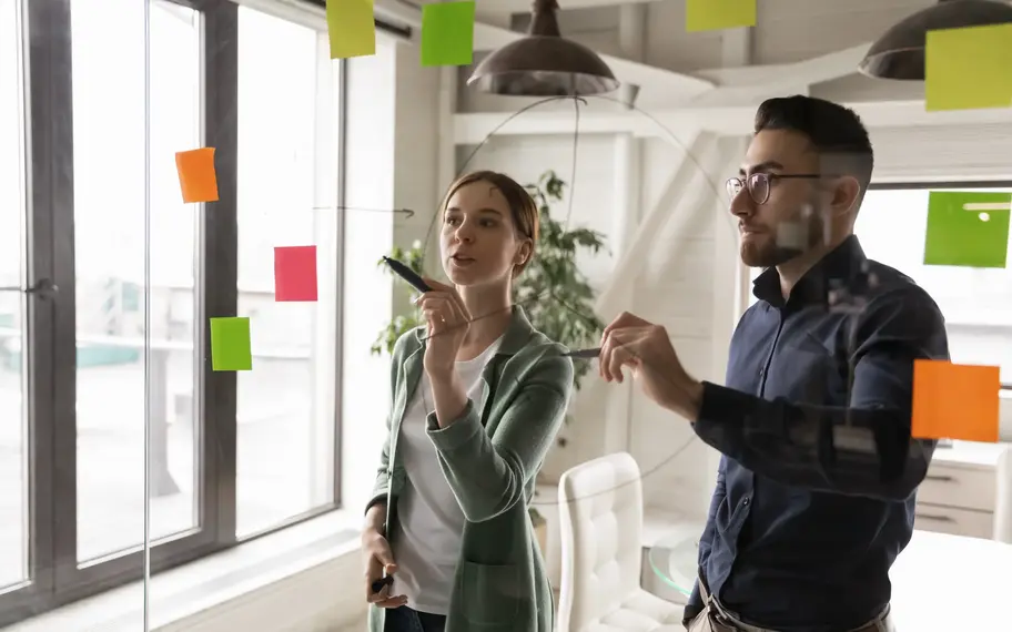 Zwei junge Personen diskutieren an einer Glaswand über Workflows diskutiert oder Arbeitsprojektprozesse: Symbolbild für Innovation und agiles Arbeiten