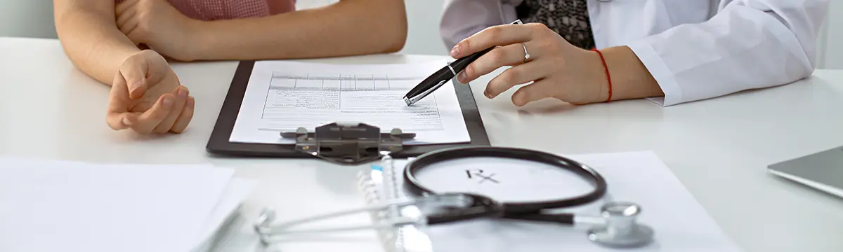 Ein Arzt und ein Patient sitzen an einem Schreibtisch und besprechen Informationen auf einem Dokument an einem Klemmbrett.