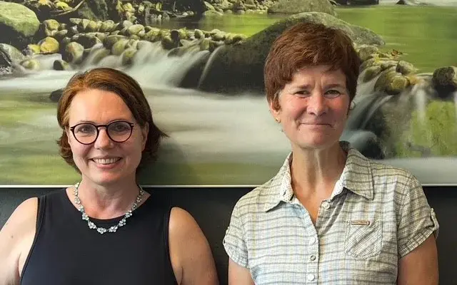 Kristine Engeleit (rechts) und Dr. Kathrin Lind Kristine Engeleit, Hausärztin, und Dr. Kathrin Lind von der AOK Niedersachsen stehen vor einem Bild eines Flusslaufs mit Steinen und grüner Vegetation