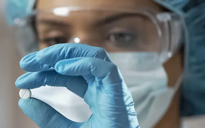 Das Bild zeigt eine Labormitarbeiterin. Sie hält eine Hand mit einem blauen Handschuh vor ihr Gesicht und prüft eine Tablette.