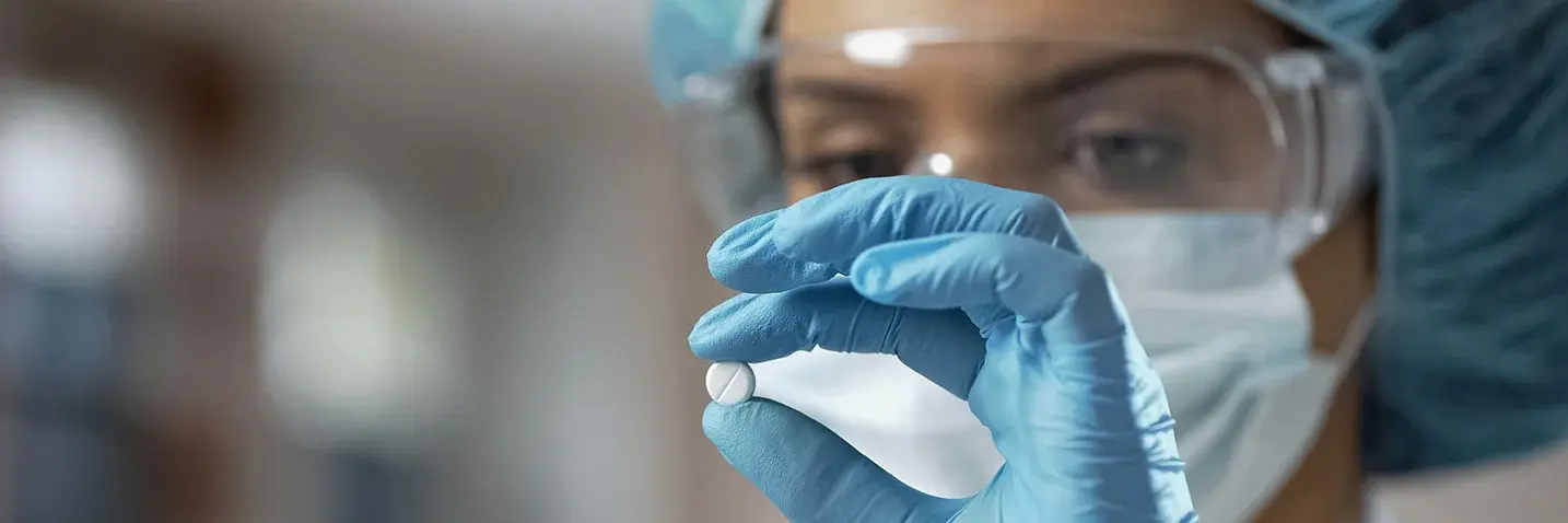 Das Bild zeigt eine Labormitarbeiterin. Sie hält eine Hand mit einem blauen Handschuh vor ihr Gesicht und prüft eine Tablette.