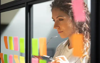 Nachdenkliche junge weibliche Teamleiterin, die sich eine Kanban/Scrum-Posts an einer Glasfenstertafel ansieht.  Symbolbild für Innovation und agiles Arbeiten.