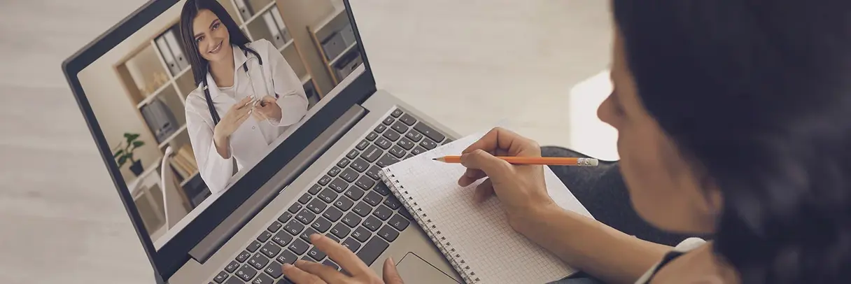 Telemedizin Frau sitzt vor einem Laptop, auf dem eine Frau mit Arztkittel zu sehen ist, und hält mit der rechten Hand einen Stift über einem Notizblock