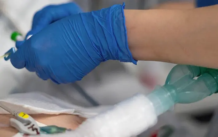 Patient wird liegt im Krankenhausbett und wird künstlich Beatmet. Eine Pflegekraft mit blauen Gummihandschuhen überprüft beim Patienten die Werte.