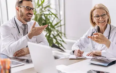 Drei Ärzte in weißen Kitteln sitzen an einem Tisch und diskutieren freundlich miteinander in einem hellen Büro