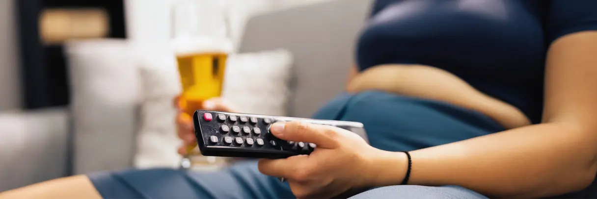 Symbol für ungesunden Lebensstil: Übergewicht, Bewegungsmangel und Alkoholkonsum Das Bild zeigt eine übergewichtige Person, die in einer Hand eine Fernbedienung und in der anderen ein Bierglas hält.