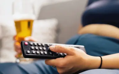 Das Bild zeigt eine übergewichtige Person, die in einer Hand eine Fernbedienung und in der anderen ein Bierglas hält.