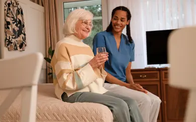 Pflegeheim: Eine Seniorin sitzt auf ihrem Bett und hält ein Glas Wasser. Eine Pflegekraft sitzt lächelnd neben ihr. 
