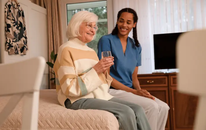 Pflegeheim: Eine Seniorin sitzt auf ihrem Bett und hält ein Glas Wasser. Eine Pflegekraft sitzt lächelnd neben ihr. 