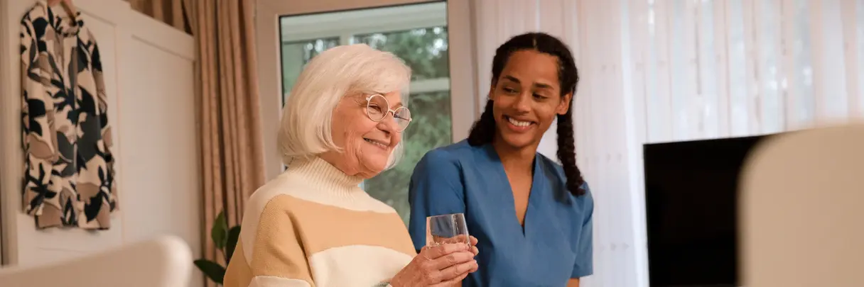 Pflegeheim: Seniorin und Pflegekraft. Pflegeheim: Eine Seniorin sitzt auf ihrem Bett und hält ein Glas Wasser. Eine Pflegekraft sitzt lächelnd neben ihr.